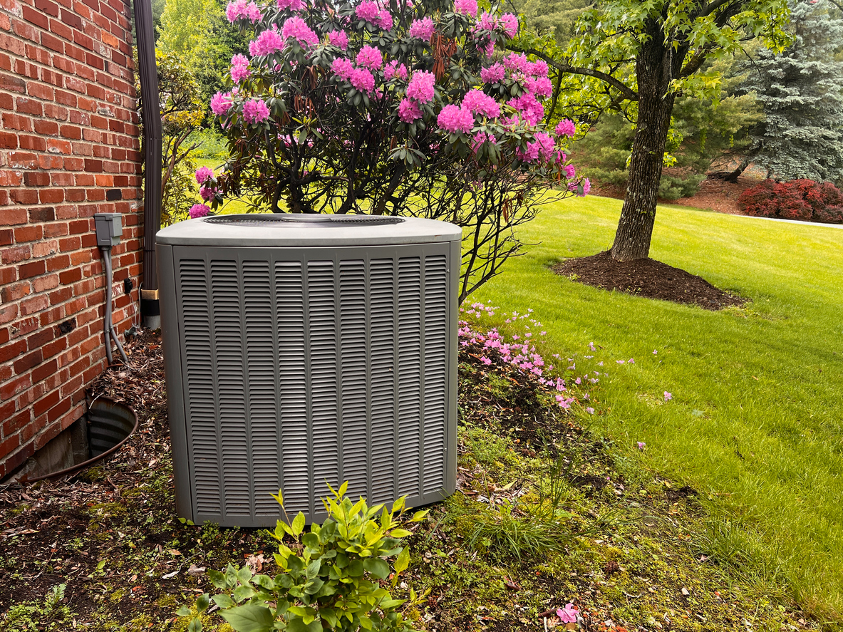 Outdoor air conditioning condenser unit beside a brick house, surrounded by blooming pink spring flowers and a green lawn.